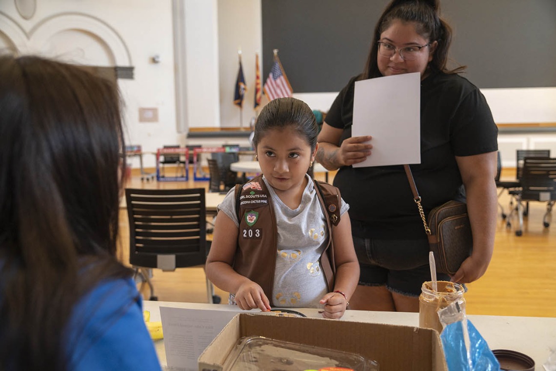 A young girl scout in uniform stands at an activity table with her mom behind her listening to a volunteer.