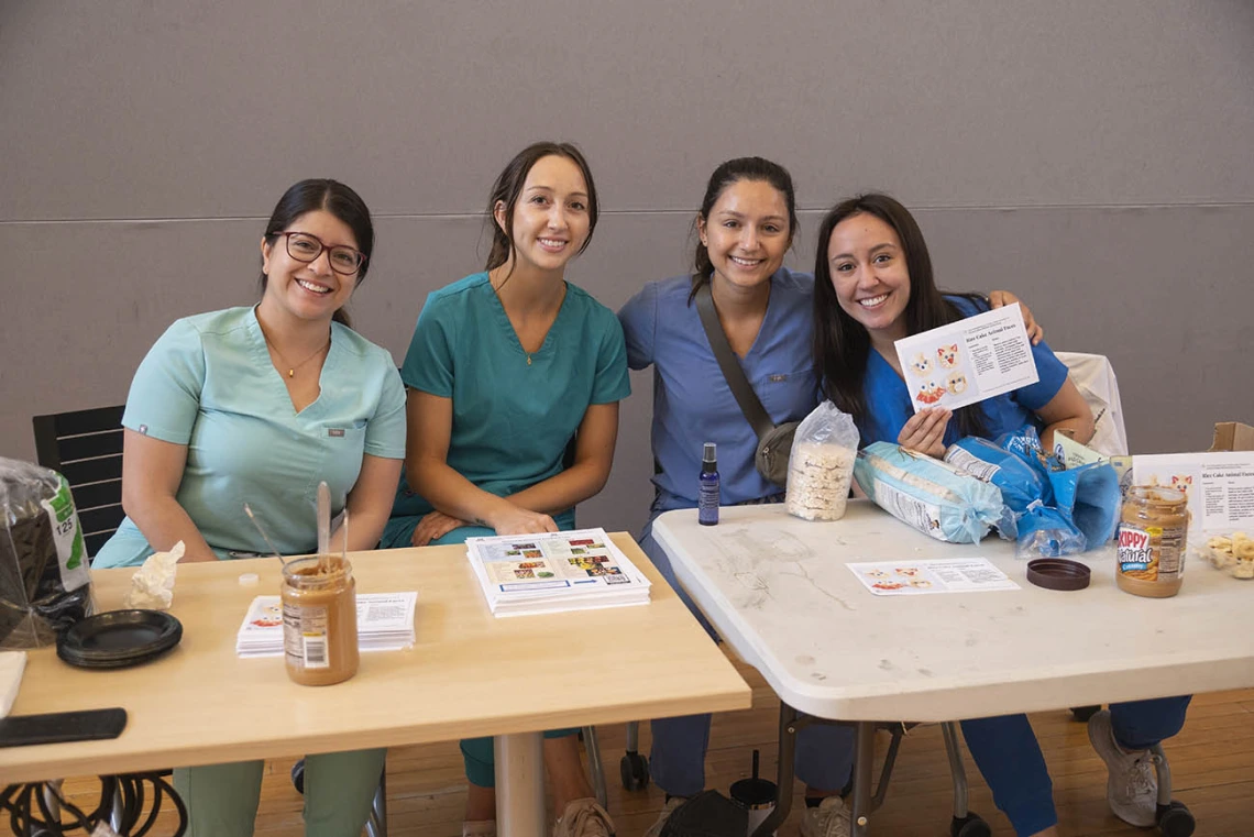 Four young women in scrubs, all medical students, sit at an activity table smiling. 