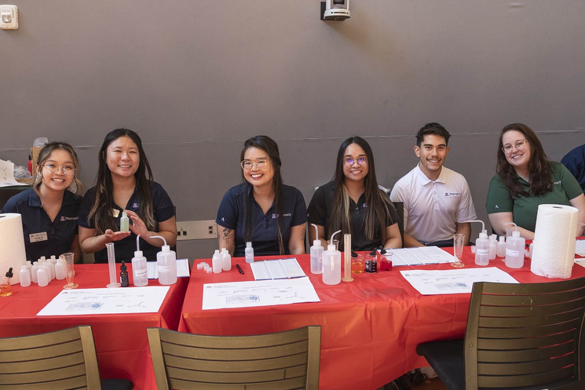 Five female and one male pharmacy students sit at an activity table smiling. 