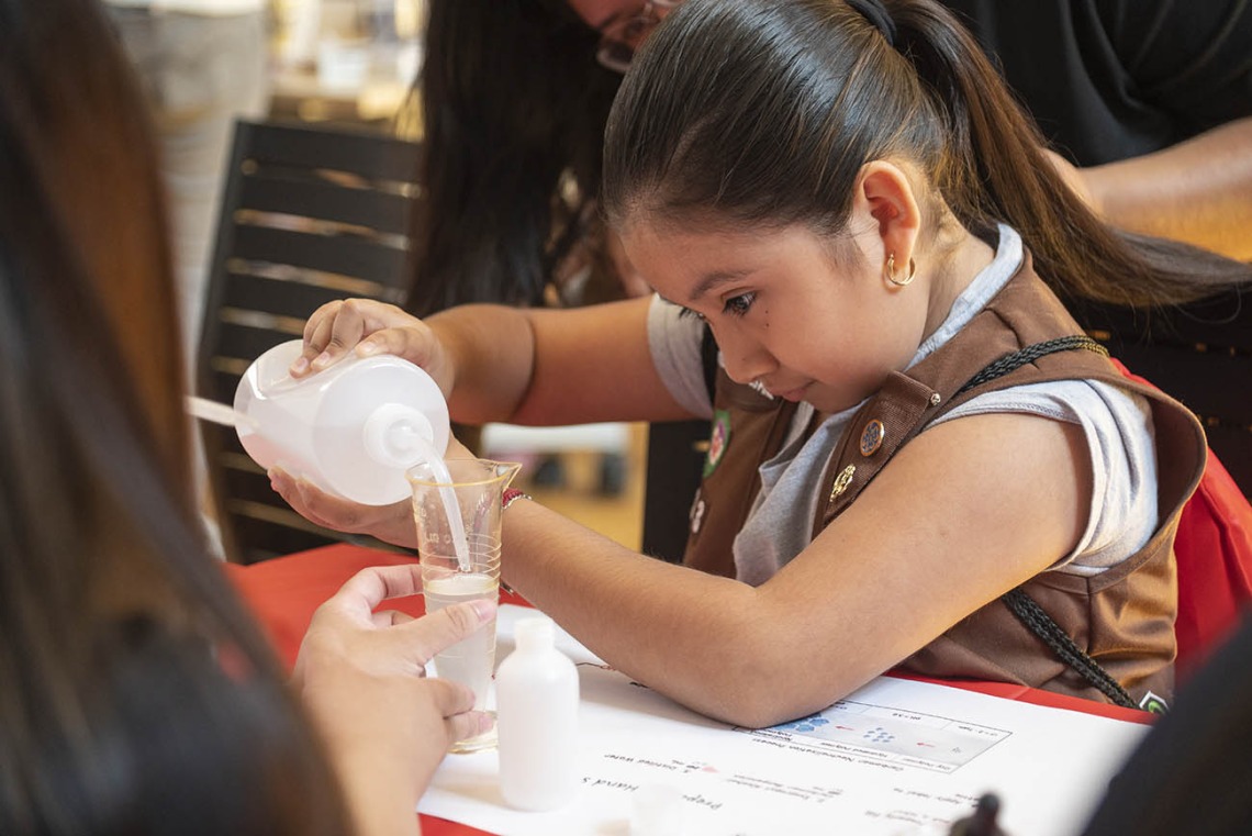 A young girl scout with dark hari in a ponytail pours liquid into a container at an activity table. 