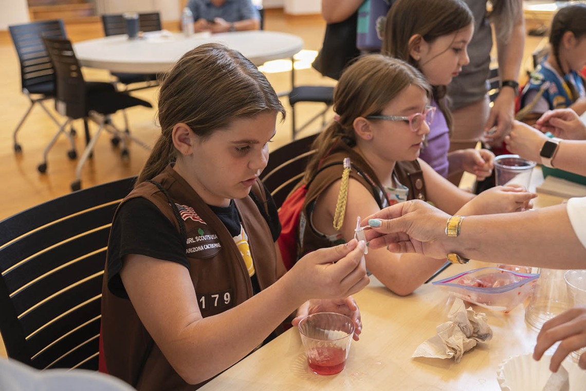 Young girls sit at an activity table with cups of liquid extracting DNA from strawberries.