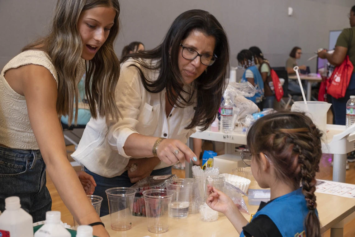 A young woman and her mother stand at an activity table helping a young girl scout.