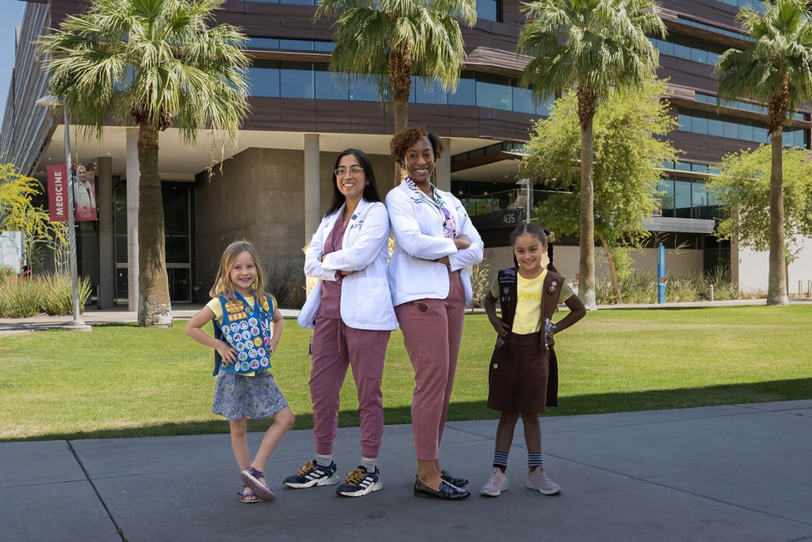 Two young girl scouts pose outside with two female medical students wearing white coats.