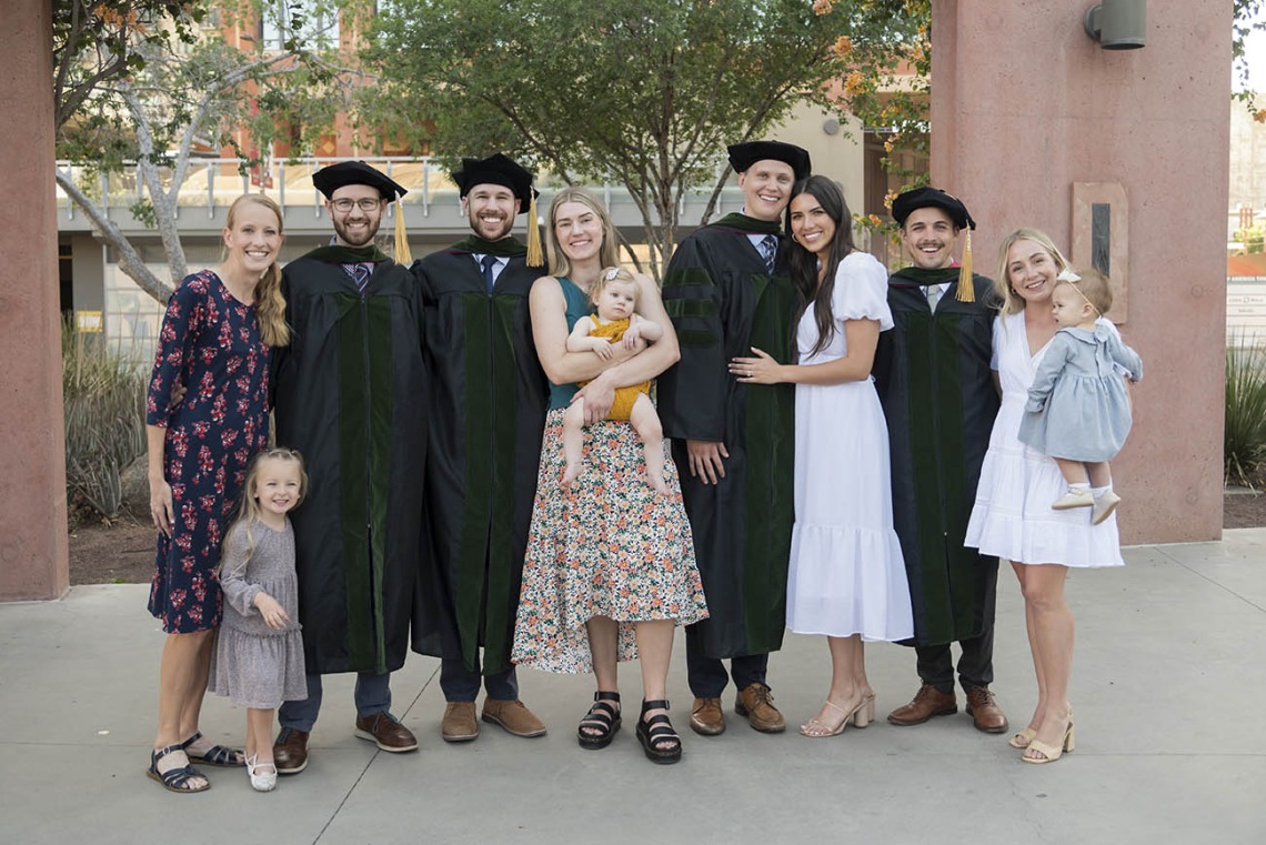 Spouses and children of UArizona College of Medicine – Phoenix class of 2022 graduates pose for a photo after the commencement ceremony.  The graduates, in black gowns from left are, Brendon Warner, MD, Jordan Yergensen, MD, Connor Swensen, MD, and Scott Litton, MD.