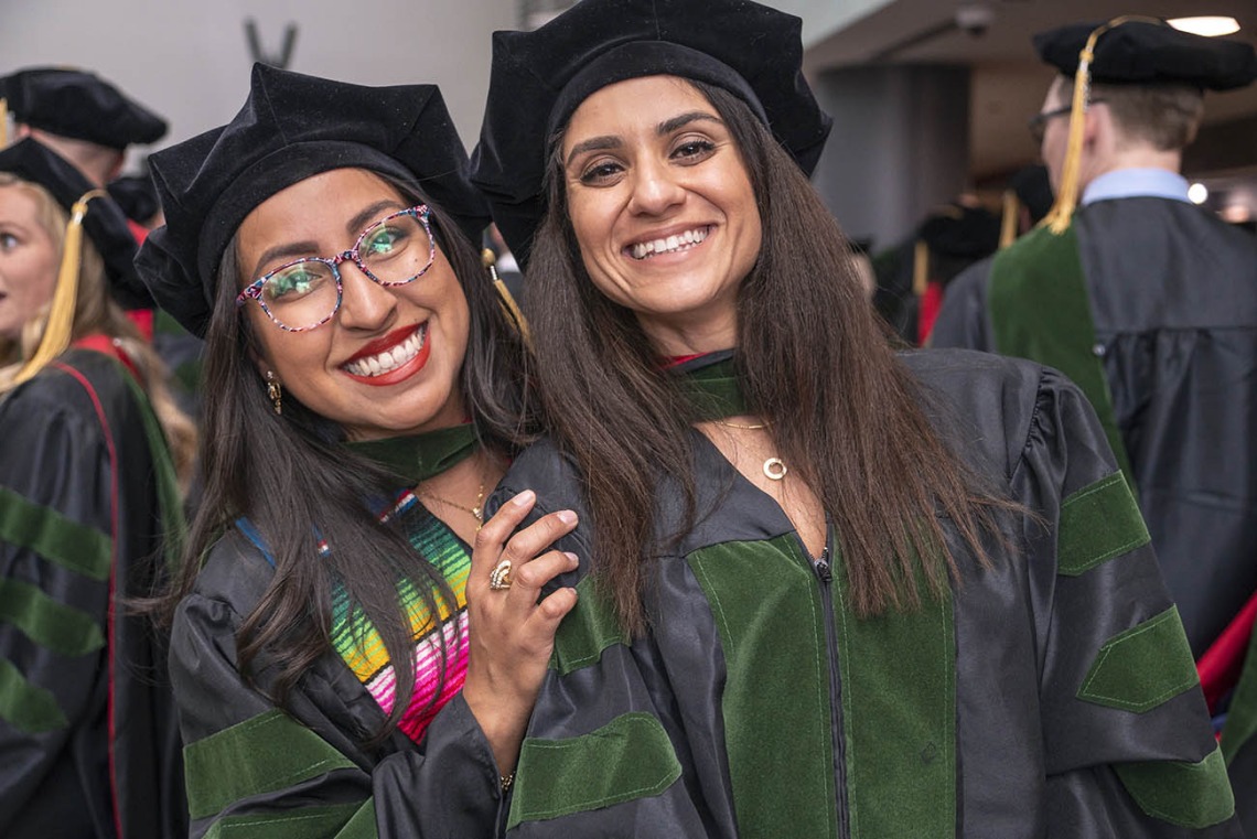 Two young women, both with long dark hair, wearing graduation regalia smile while standing closely together. 