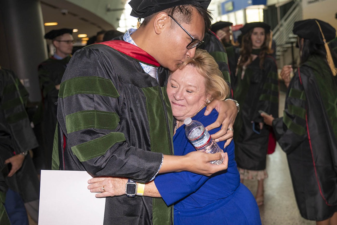 A tall young brown-skinned man wearing graduation regalia hugs a shorter, older blonde woman. 