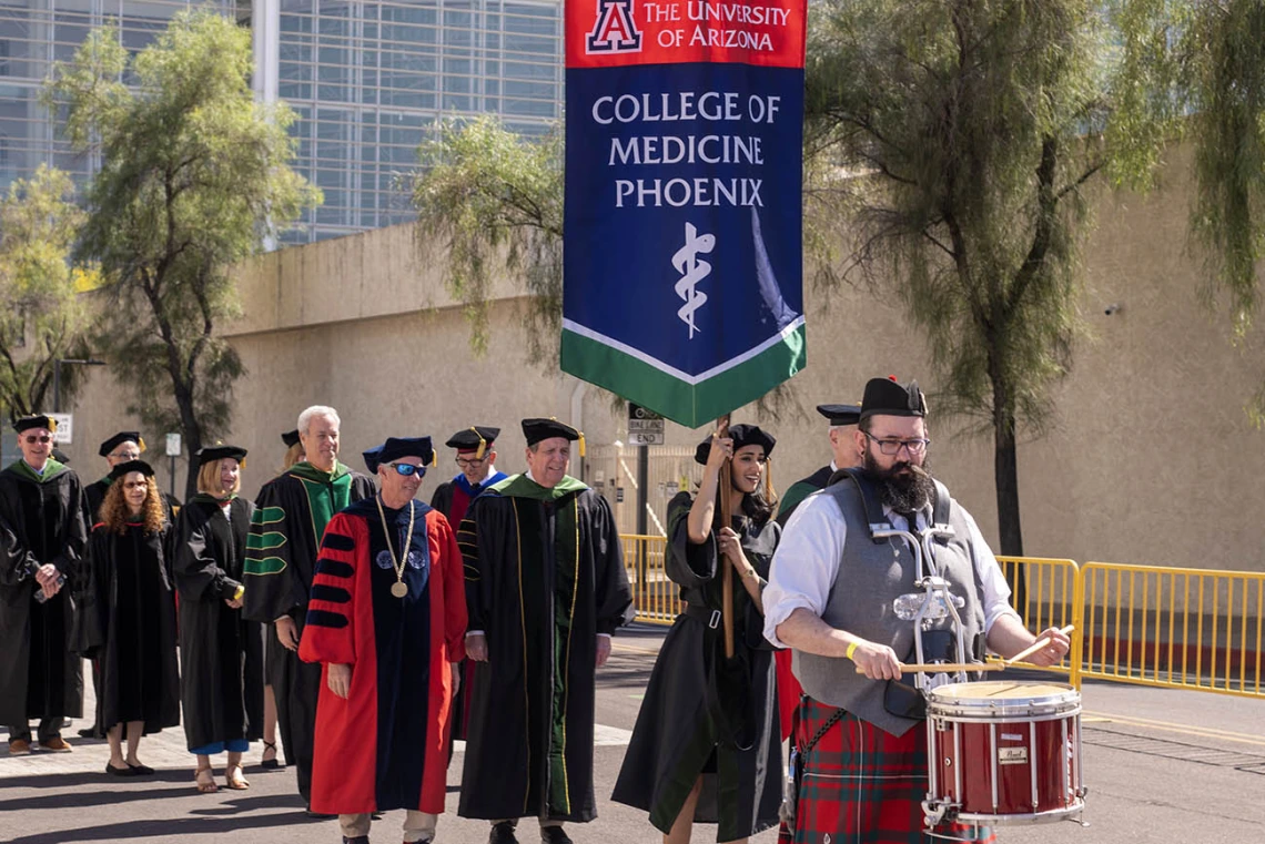 A bearded man wearing a kilt and playing a drum leads a procession of of people wearing graduation regalia down the street.