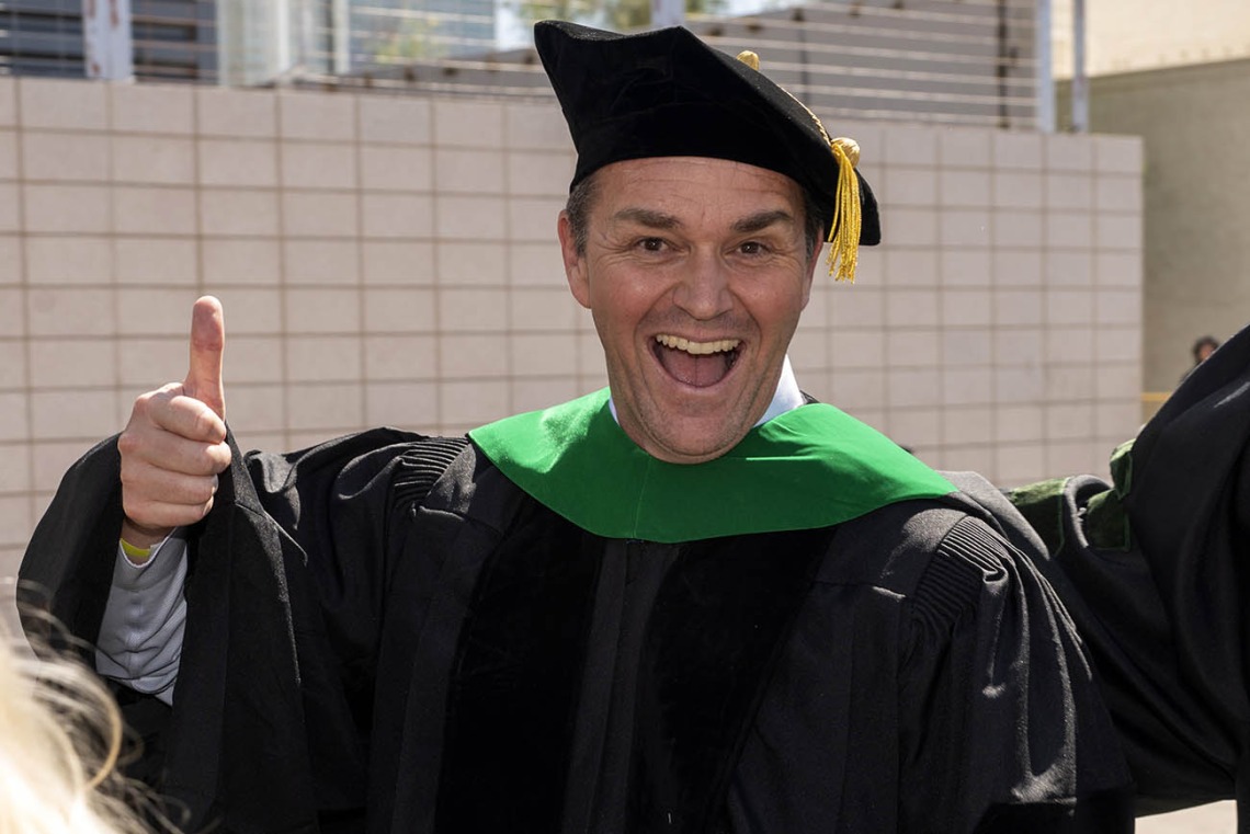 A middle-aged man wearing graduation regalia smiles and gives a thumbs-up.