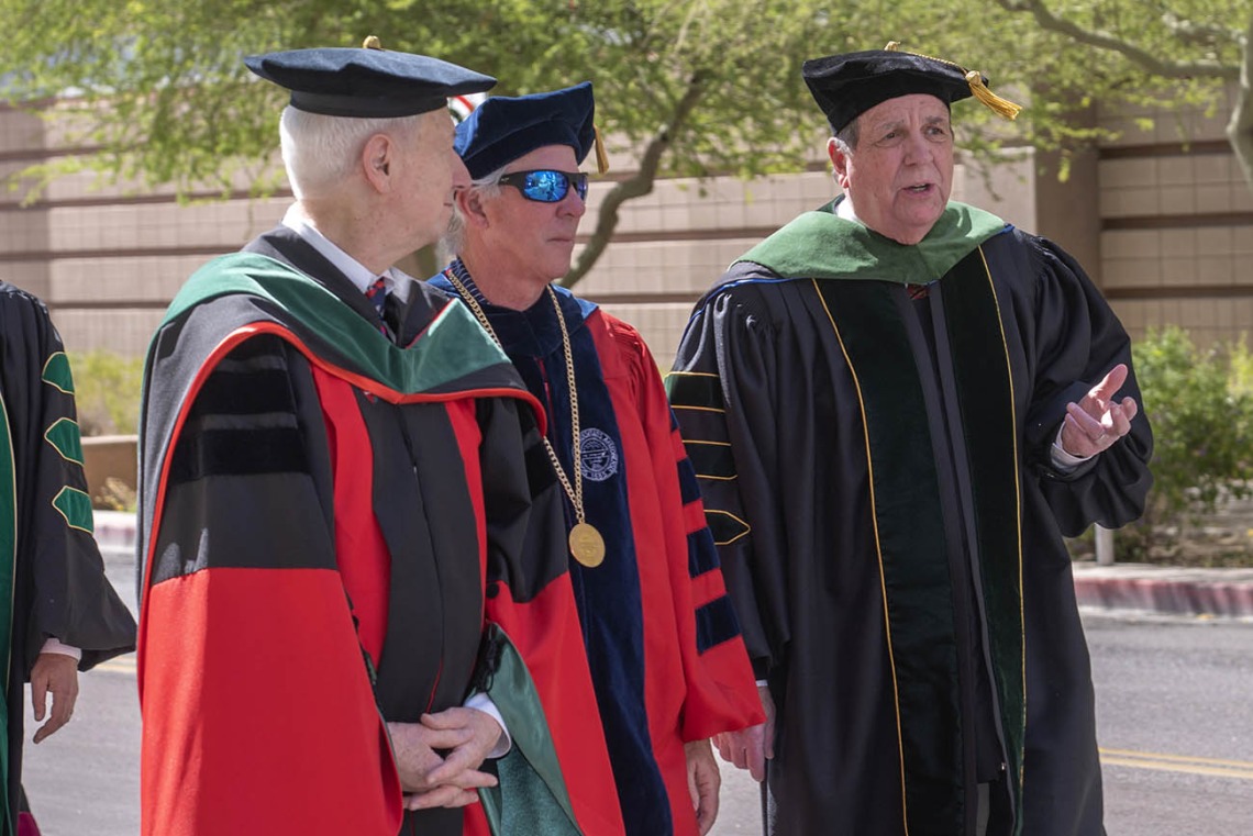 Three older men wearing graduation regalia chat while walking outside side-by-side.