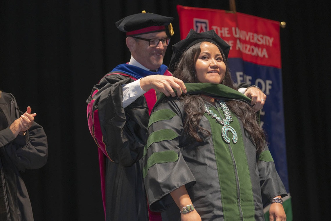 A young Native American woman dressed in graduation cap and gown has a sash placed over her shoulders by a male faculty member. 