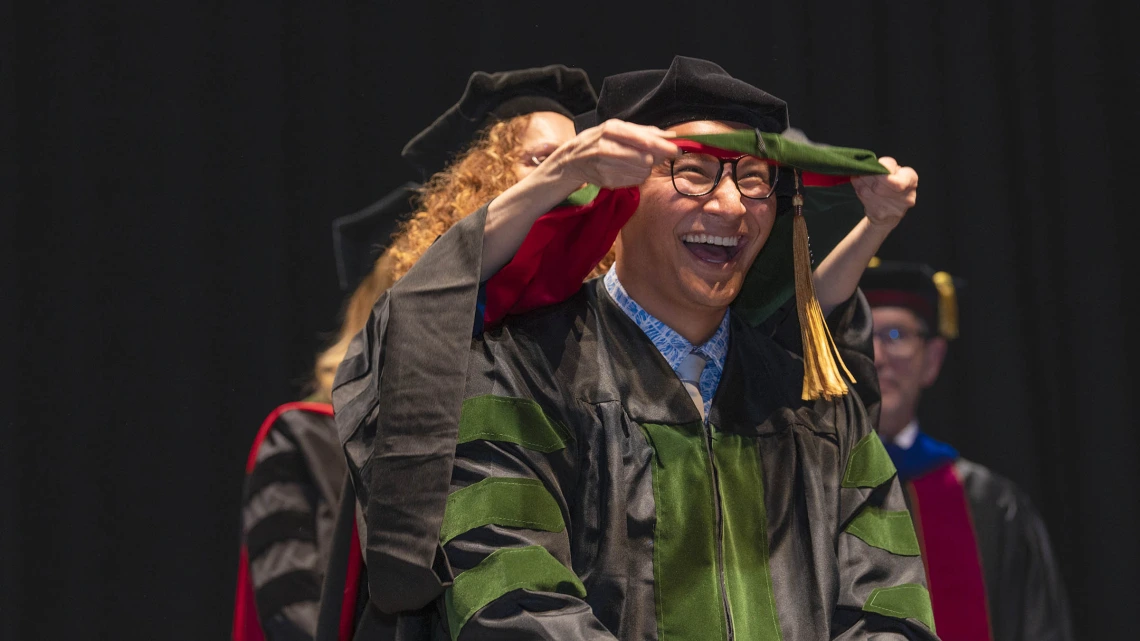 A young man with a big smile is wearing a graduation cap and gown has a sash placed over his shoulders. 