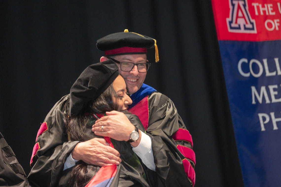 A young woman wearing graduation regalia hugs a male faculty member wearing regalia. 