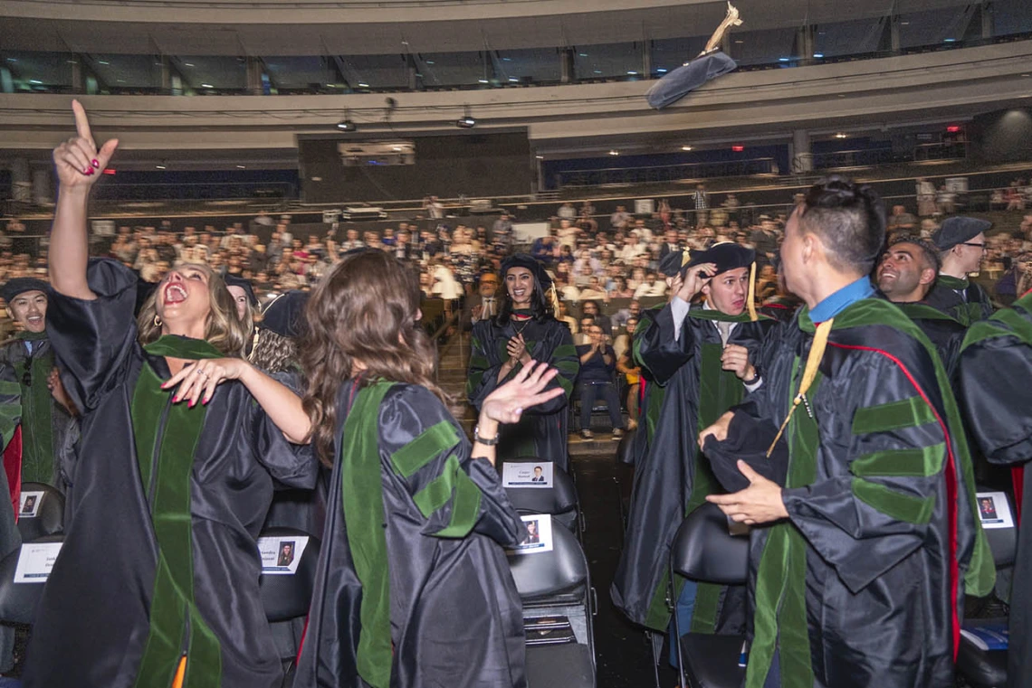 A large group of medical students wearing graduation regalia celebrate by throwing their hats into the air.