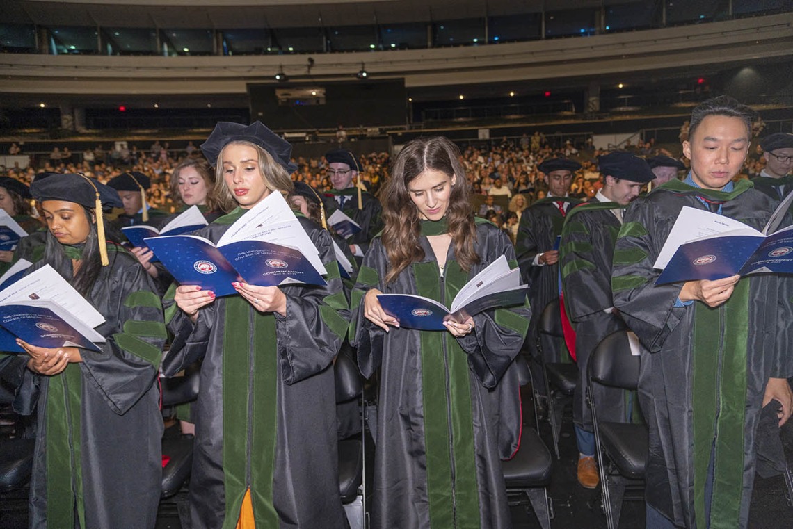 A large group of medical students wearing graduation regalia read from a pamphlet at their graduation.