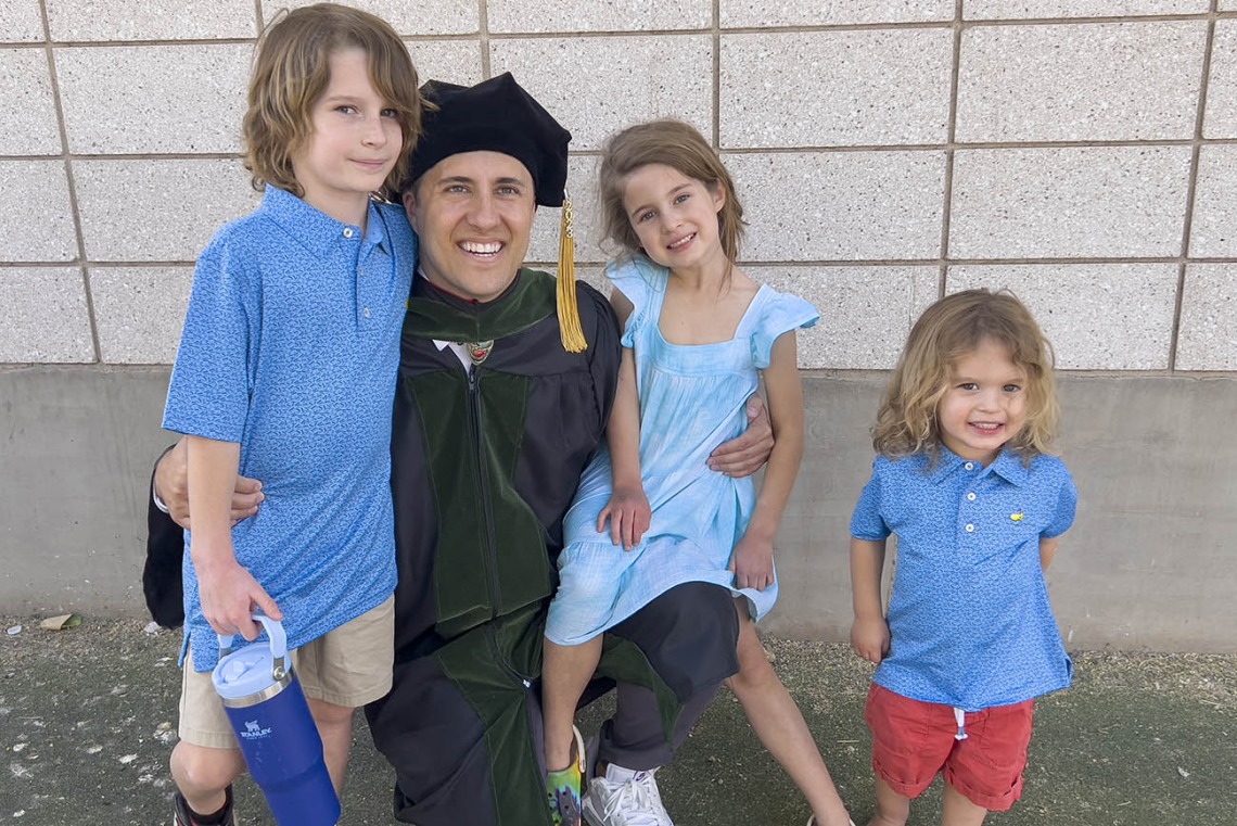 A light-skinned young man in graduation regalia kneels down and is surrounded by his three young children. 