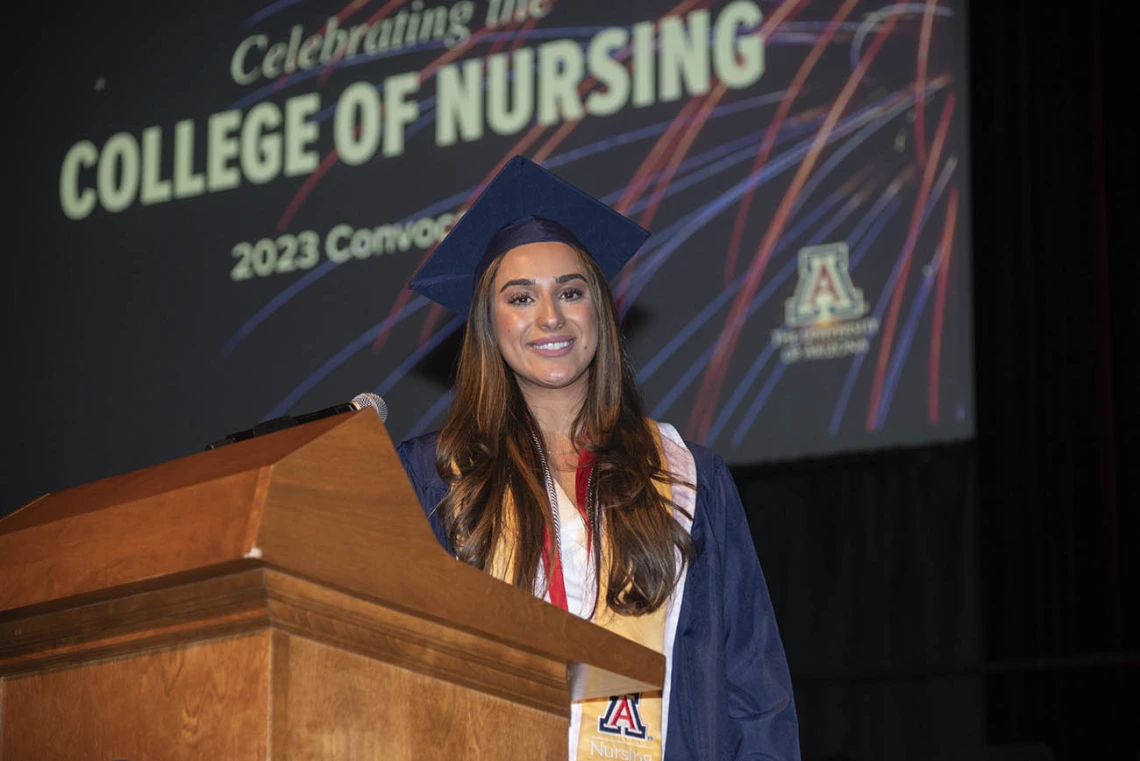 A young woman with long brown hair and dressed in cap and gown stands at a podium in Centennial Hall.