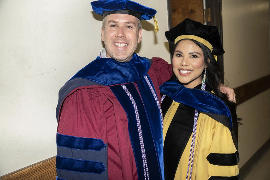 A male professor and a female professor dressed in graduation regalia backstage in Centennial Hall.