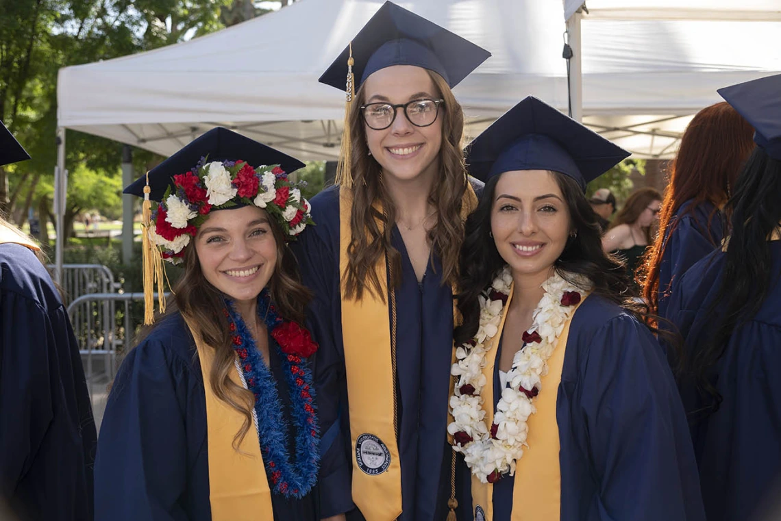 Three smiling women dressed in graduation regalia stand outside after their convocation. 