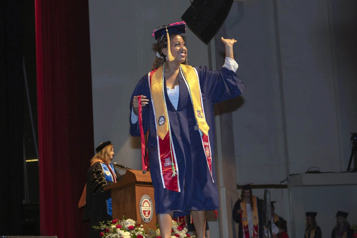 A young woman dressed in graduation regalia waves as she walks across the stage at Centennial Hall.