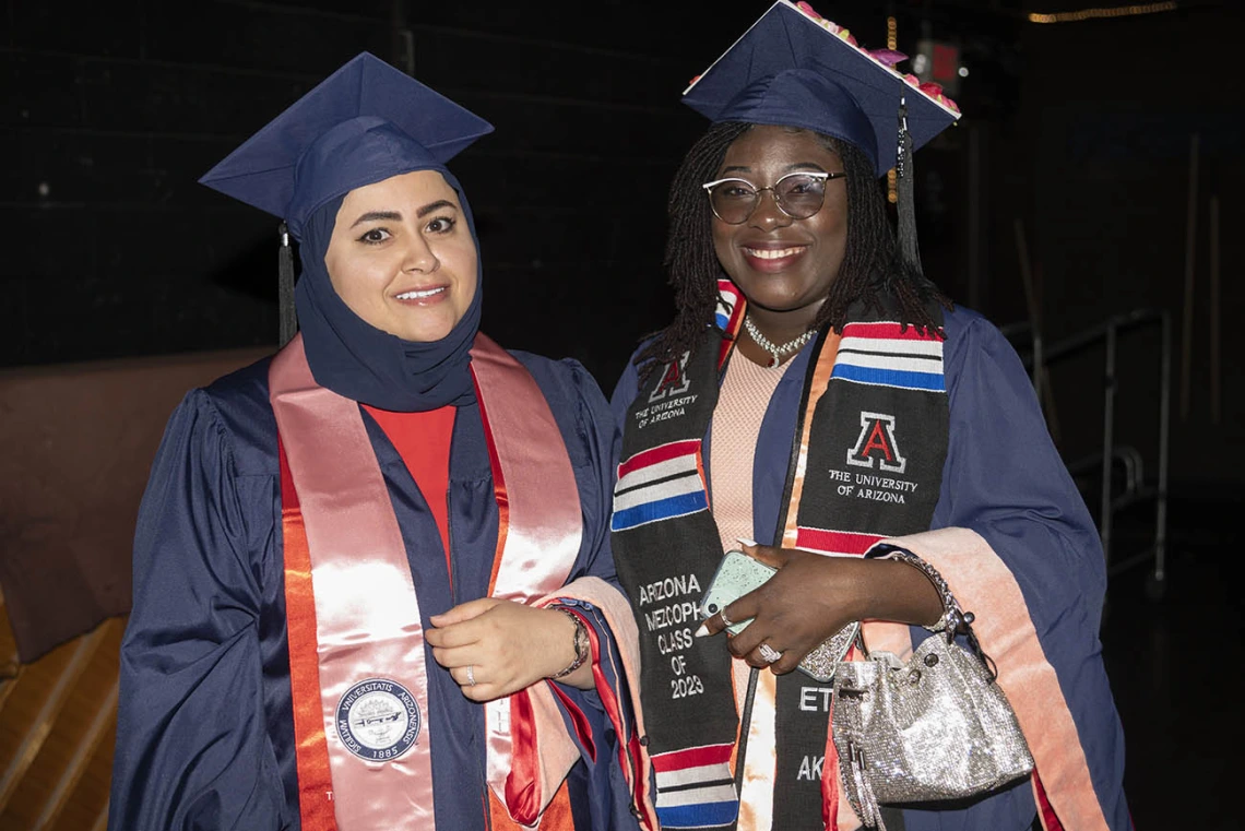 Two young women stand side-by-side wearing graduation regalia smiling. 