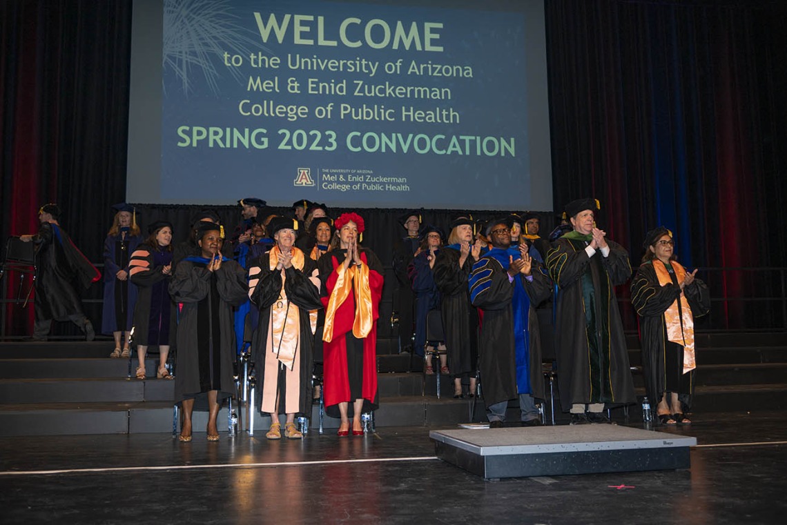 A large group of faculty members in graduation regalia stand on a stage under a large screen reading welcome to spring convocation.