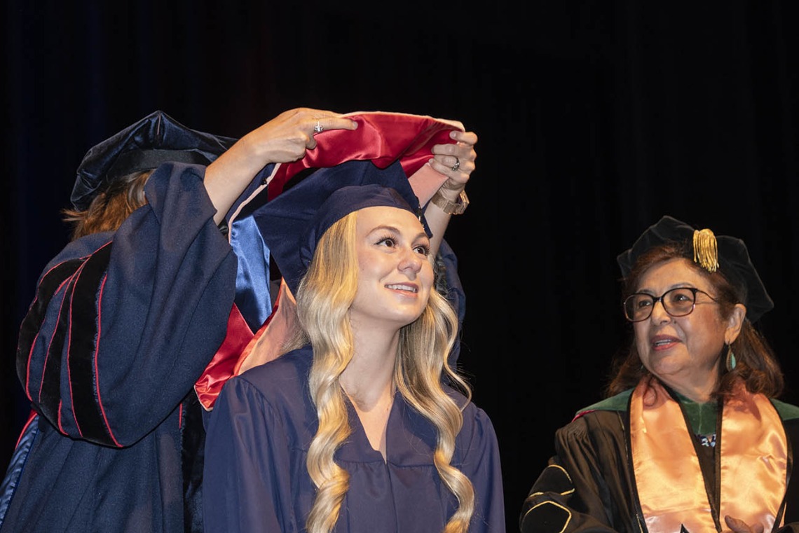 A smiling young woman with long blonde hair wearing a graduation cap and gown looks up as a professor places a sash over her shoulders. 