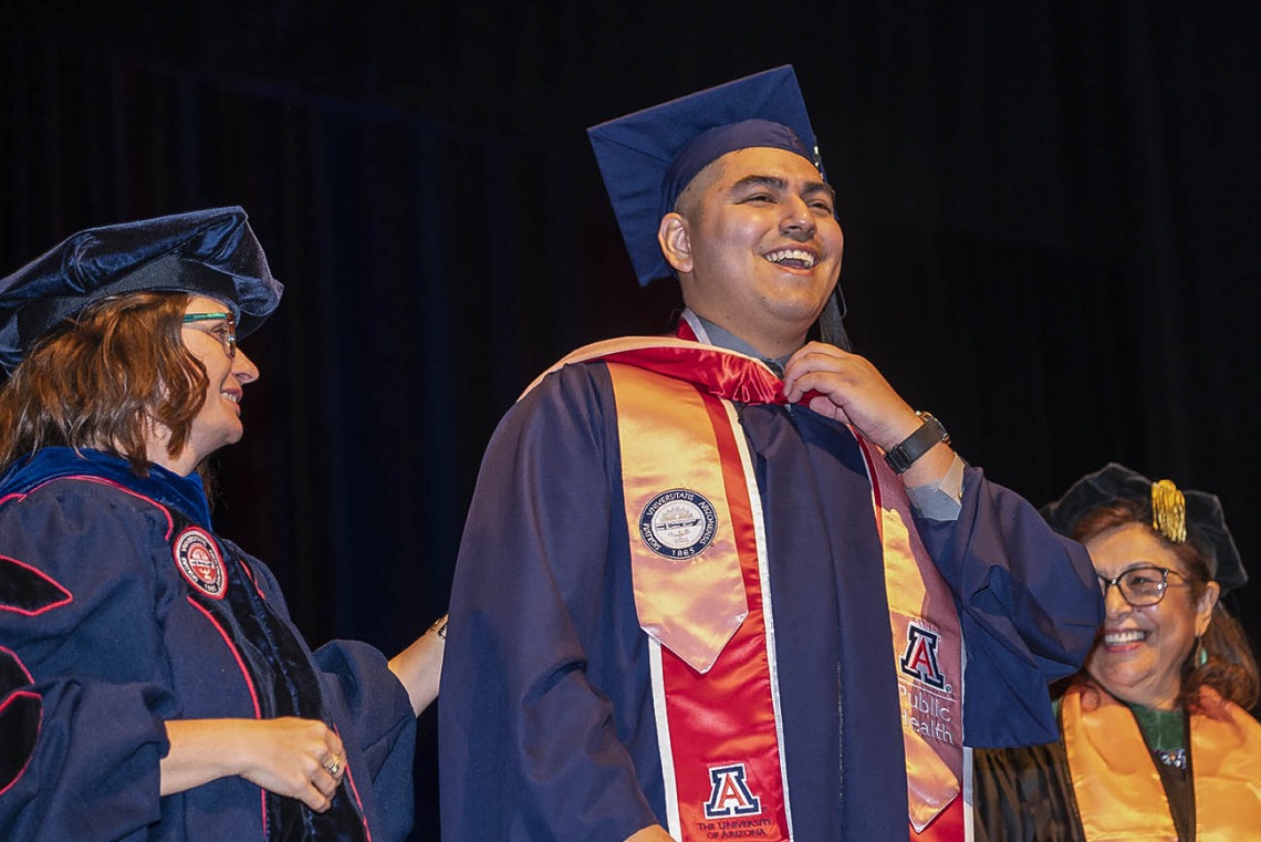 A smiling young man wearing a graduation cap and gown stands in front of two femal professors, also in graduation regalia. 