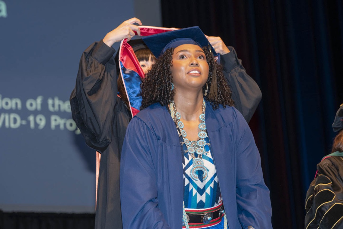 A young brown-skinned woman in graduation regalia stands smiling as a professor standing behind her places a sash over her shoulders. 