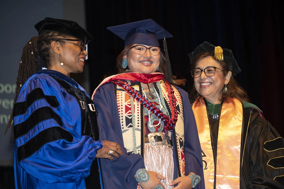 A smiling young woman wearing traditional Native American clothing under her graduation cap and gown stands with two professors.