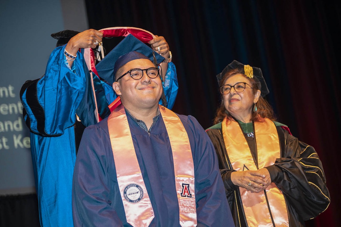 A smiling young man wearing a graduation cap and gown has a sash placed over his shoulders while the dean of the college looks on. 