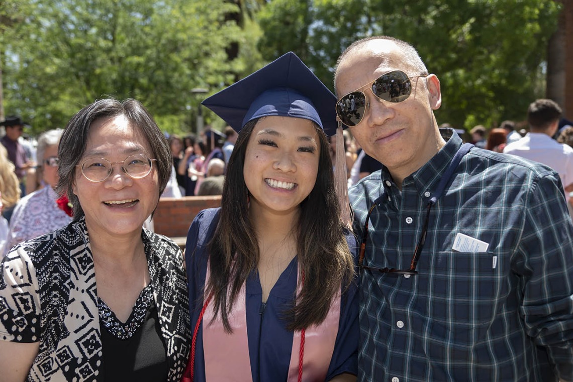 A smiling young woman in graduation cap and gown stands between her parents.
