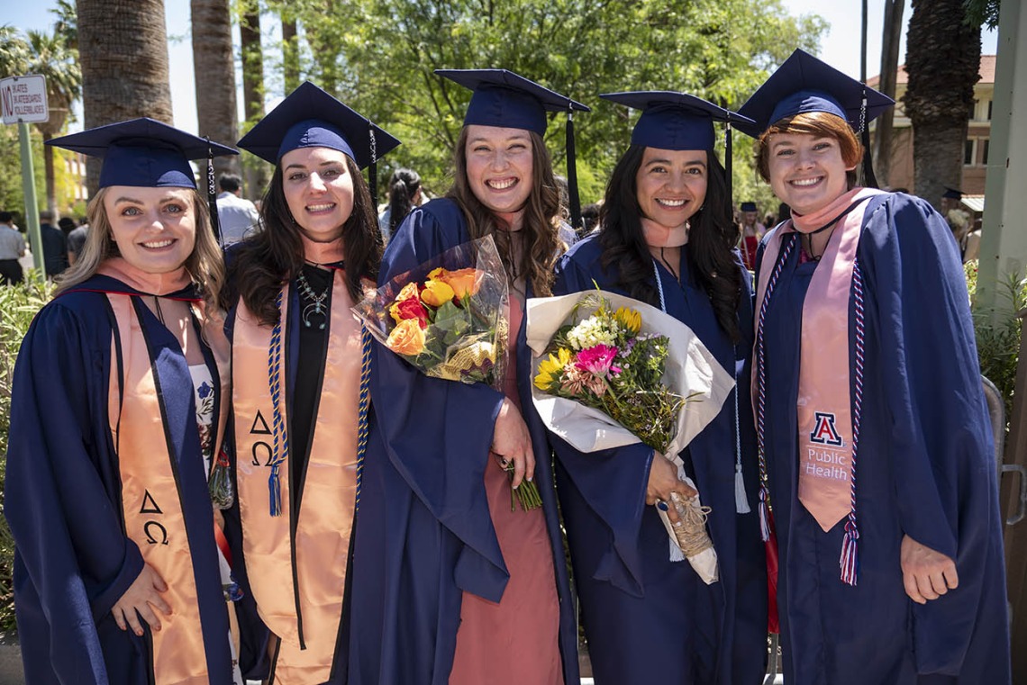 Five young women in graduation regalia stand side-by-side outside smiling, two are holding flowers. 