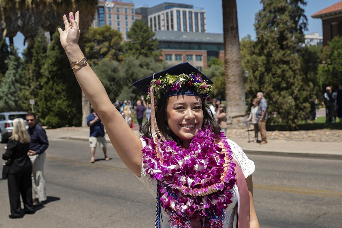 A smiling young woman wearing a graduation cap with several flower garlands around her neck waves while standing outside. 