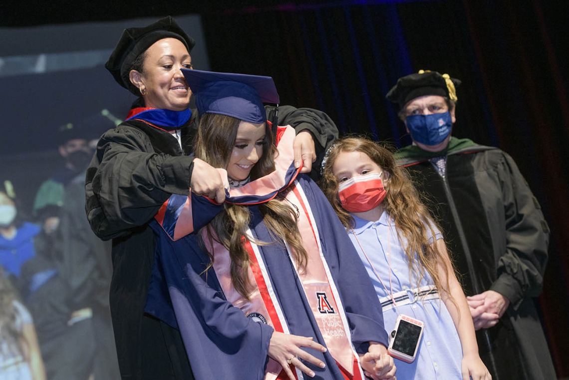 Gliney Clarissa Rios, MPH, accompanied by her daughter Mia, is hooded during the 2022 Mel and Enid Zuckerman College of Public Health spring convocation. 