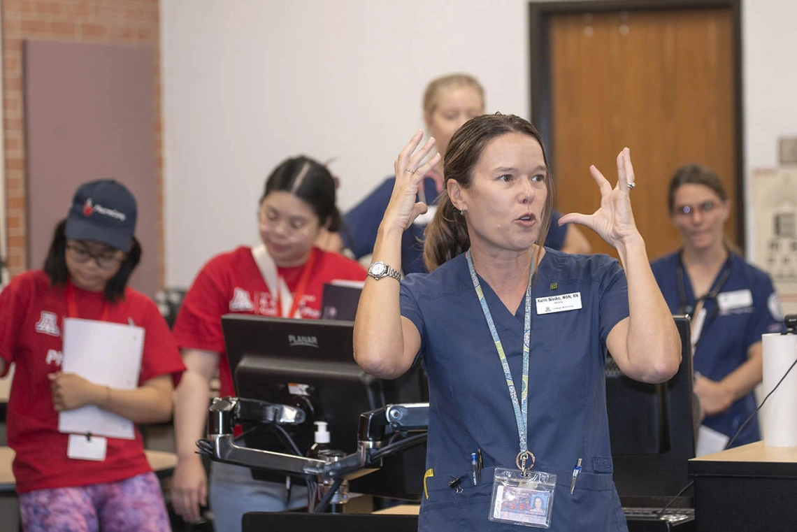 A middle-aged light skinned woman in scrubs motions with her hands as she is talking to studetns.