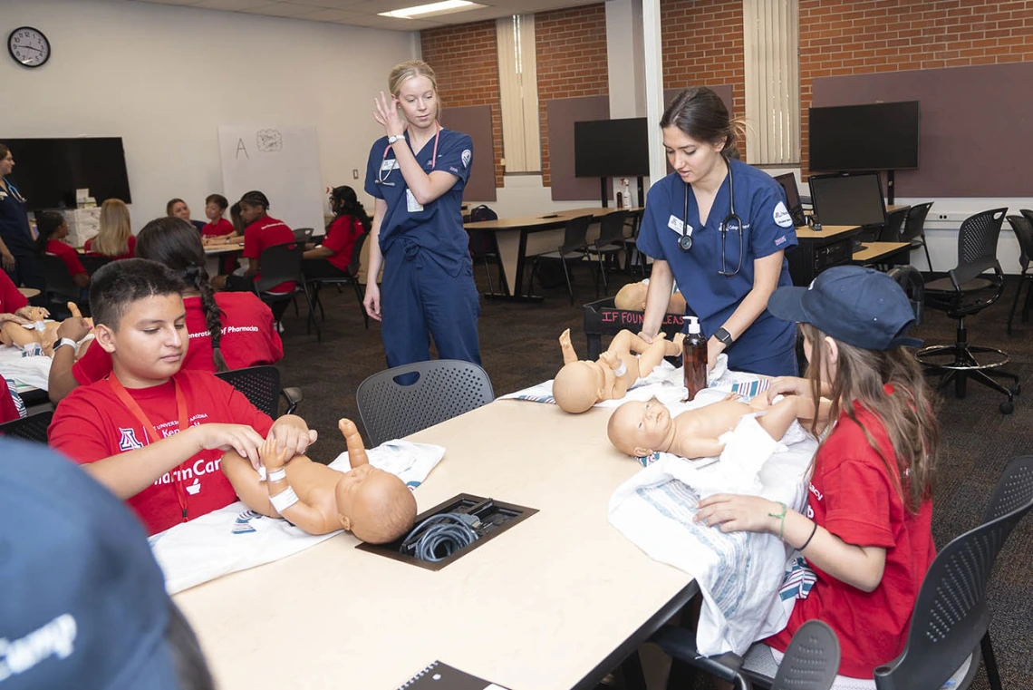 A nursing student in blue scrubs shows several young students how to change a diaper on a baby manikin.