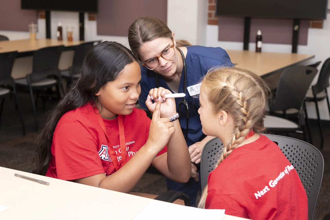 A young nursing student in blue scrubs watches as two girls check each other's pupils with a flashlight.