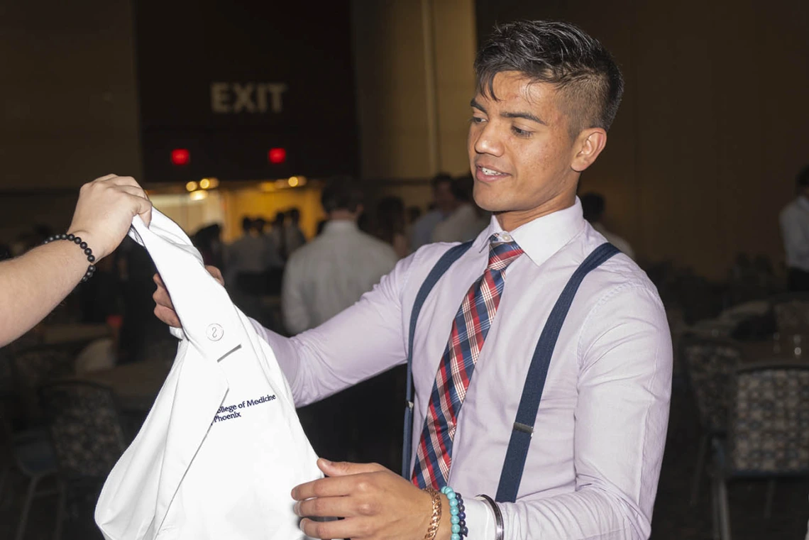 A young man with short dark hair and light brown skin wearing a button-up shirt, tie and suspenders reaches for a white coat. 