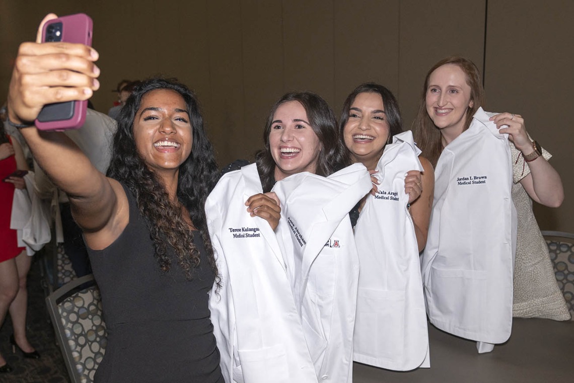 A group of four young women smile as they hold up their medical school white coats as one of them takes a selfie of the group. 