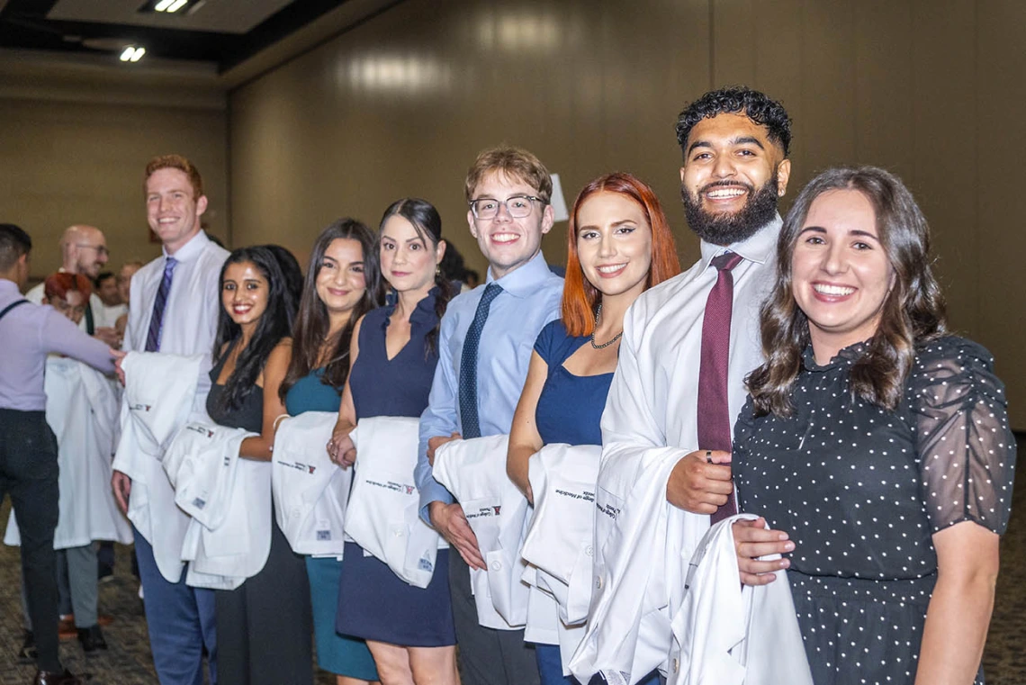 Several medical school students stand in a line smiling with white coats over their arms. 