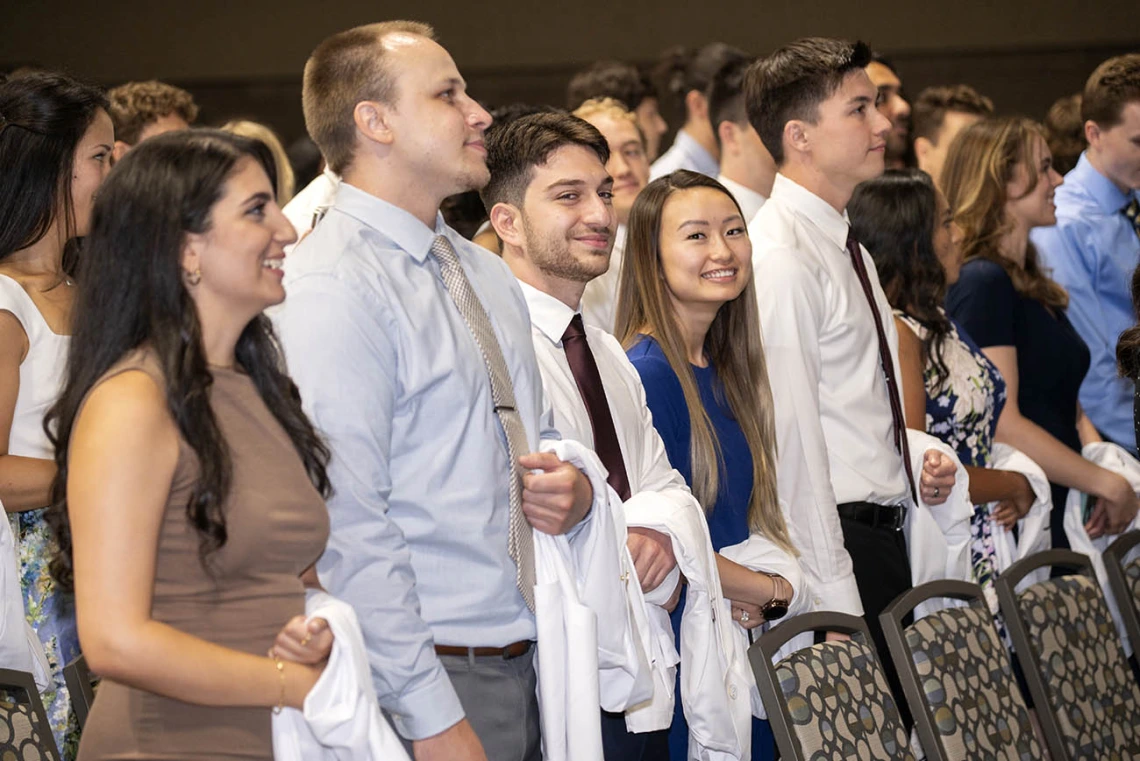 Many medical school students with white coats over their arms stand in front of chairs preparing to go onto stage.