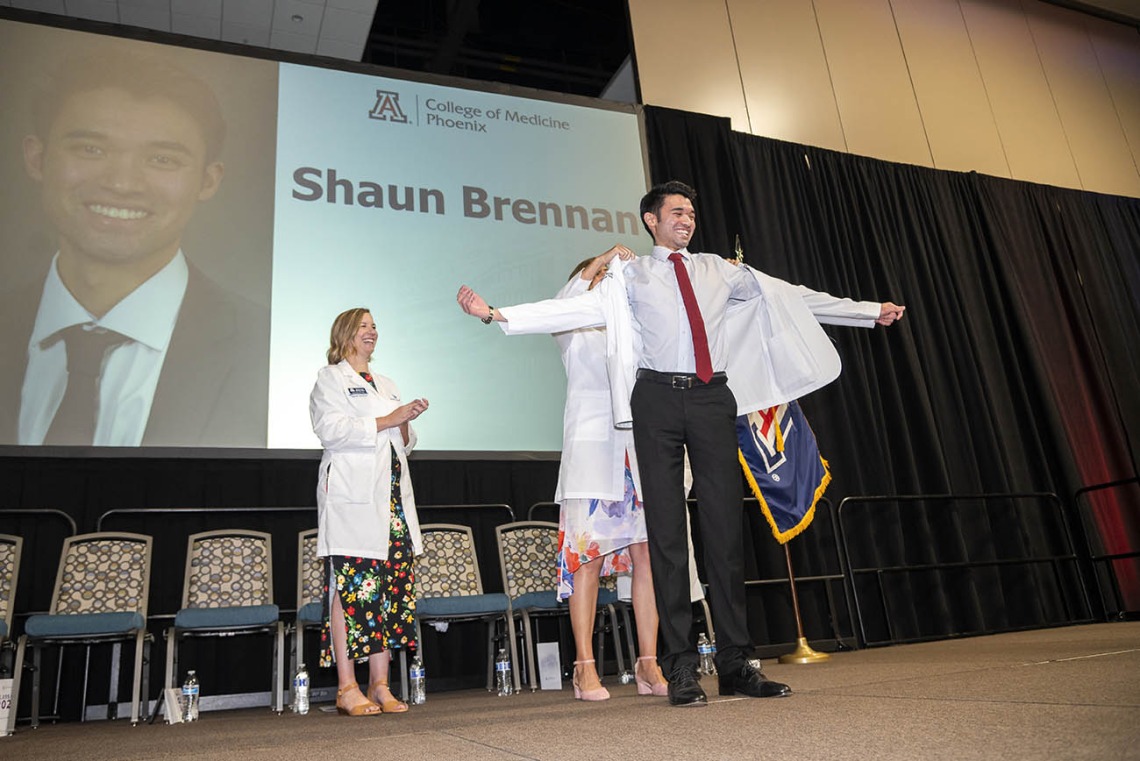 A young man stands on a stage smiling with his arms outstreatched as a faculty member helps put a white coat on him.