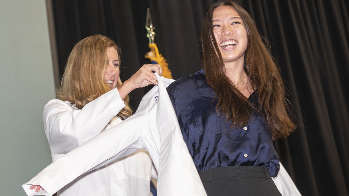 A white woman in a doctor's coat helps a young Asian woman with a big smile put on a white doctor's coat. 