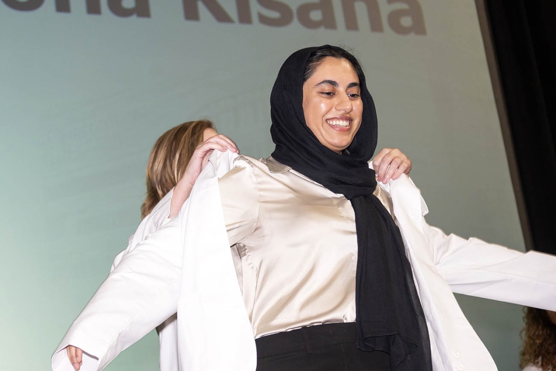 A young woman with her hair covered by a dark scarf smiles as she has her white coat placed on her. 