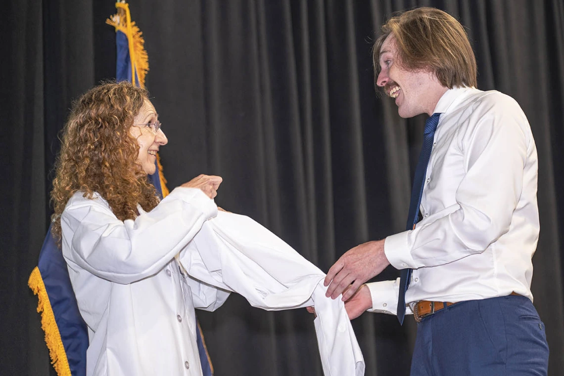 A white female doctor helps a young male medical student with light skin and a mustache put on his white coat. 