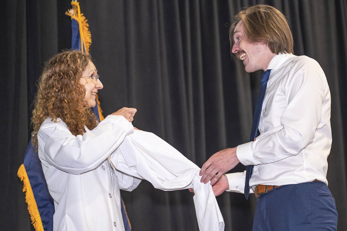 A white female doctor helps a young male medical student with light skin and a mustache put on his white coat. 