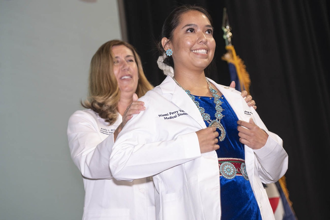 A white female doctor helps a young female medical student wearing a traditional Native American necklace and belt put on her white coat. 