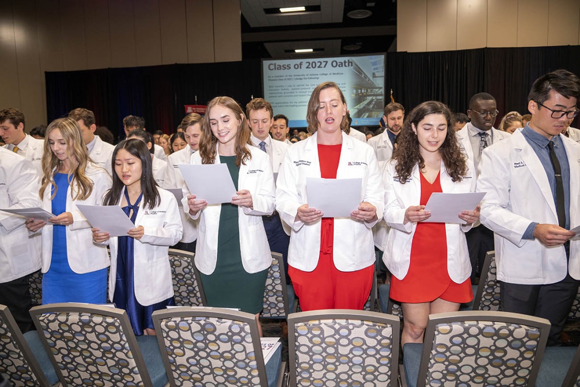 A large group of medical students wearing white coats and holding papers recites their oath.