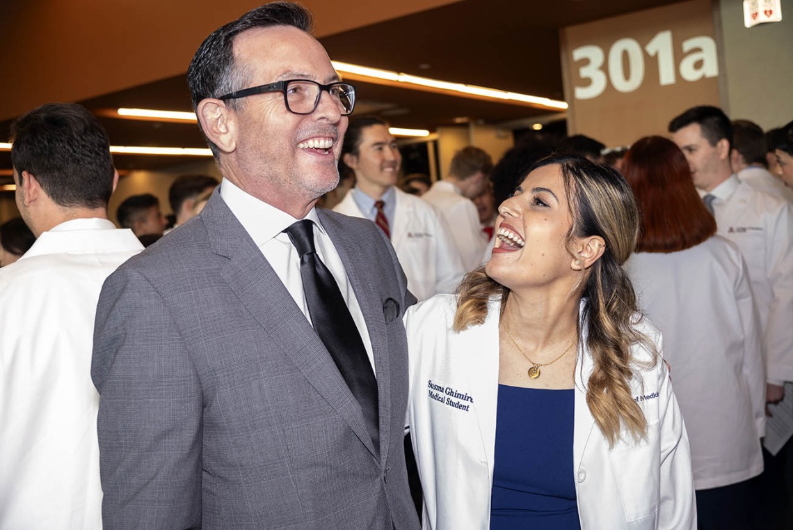 A tall white man in a suit laughs as he stands next to a shorter young woman in a white medical coat who is also laughing. 