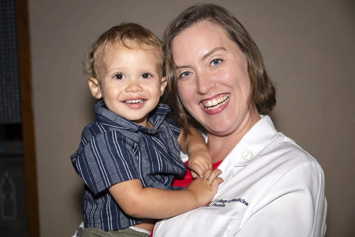 A young white woman in a medical coat smiles as she holds a little boy. Both are smiling. 